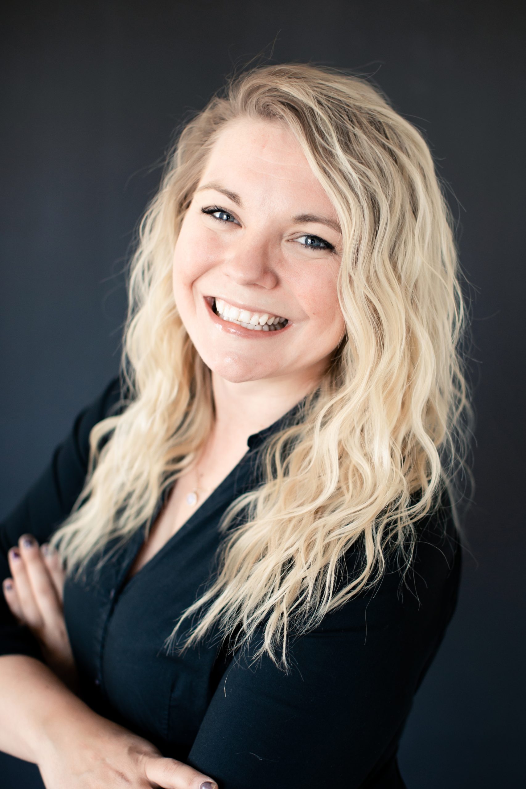 PR & Marketing Director Amy Lhotka, with wavy blonde hair, smiles confidently in a black top with arms crossed against a dark background.