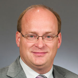 A middle-aged man with glasses and short light brown hair wearing a gray suit, white shirt, and striped tie, posed against a plain grey background. A middle-aged man with glasses and short light brown hair wearing a gray suit, white shirt, and striped tie, posed against a plain grey background.