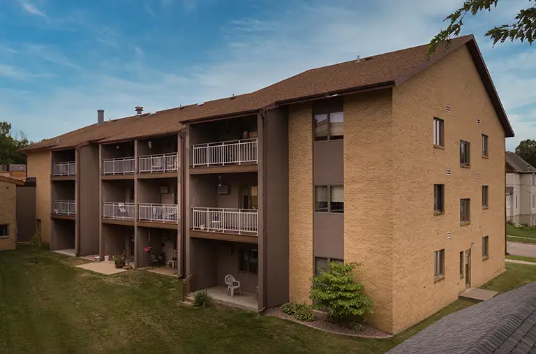 Three-story apartment building with brick exterior, balconies on the second and third floors, and a grassy yard in front under a partly cloudy sky. Three-story apartment building with brick exterior, balconies on the second and third floors, and a grassy yard in front under a partly cloudy sky.