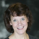 Woman with short, curly brown hair smiling at the camera, wearing a dark top against a blurred, dark background at St. Francis Outpatient Therapy in Minnesota. Woman with short, curly brown hair smiling at the camera, wearing a dark top against a blurred, dark background at St. Francis Outpatient Therapy in Minnesota.