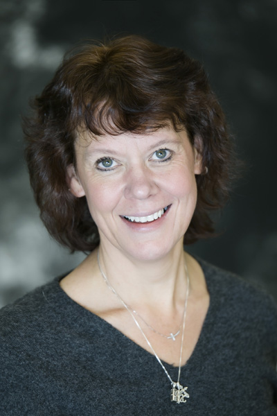 Cami Peterson-DeVries, wearing a grey sweater and two silver necklaces, smiles at the camera with short brown hair against a dark, blurred background—perfect for celebrating a work anniversary.