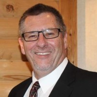 A middle-aged man with glasses, short dark hair, and a trimmed beard is smiling while wearing a suit and tie. Wooden paneling is visible in the background at St. Francis Health Services in Minnesota. A middle-aged man with glasses, short dark hair, and a trimmed beard is smiling while wearing a suit and tie. Wooden paneling is visible in the background at St. Francis Health Services in Minnesota.