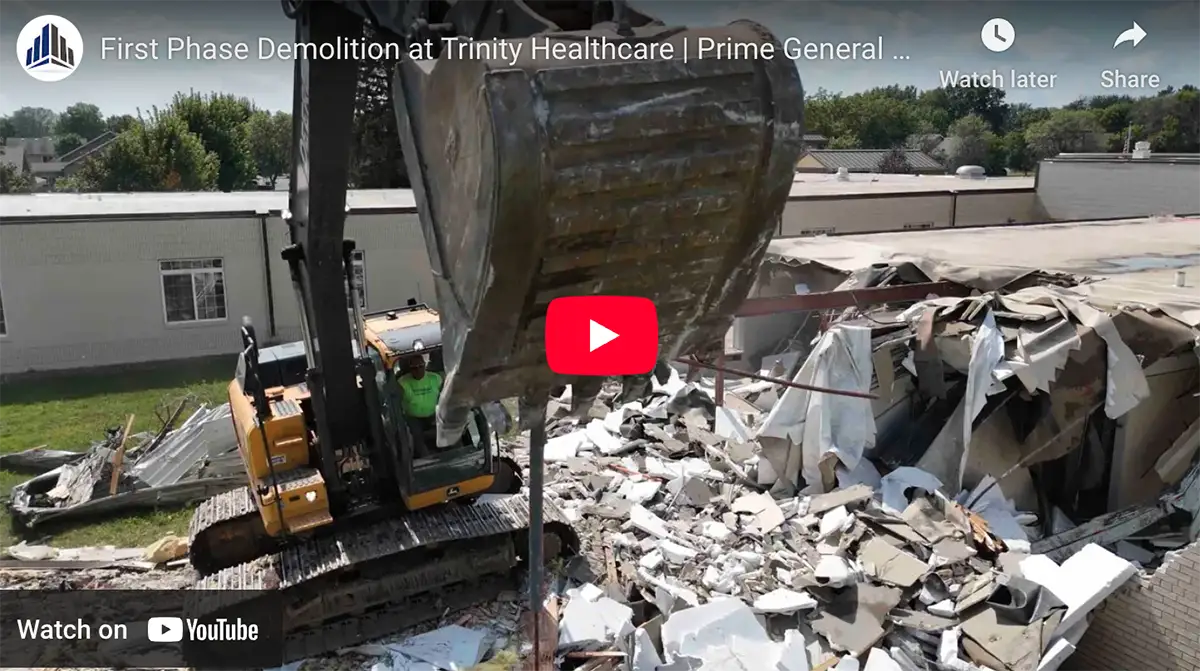 An excavator demolishes a building at Trinity Healthcare, with debris and rubble scattered around the site. Catch the current progress as demolition enters its first phase.