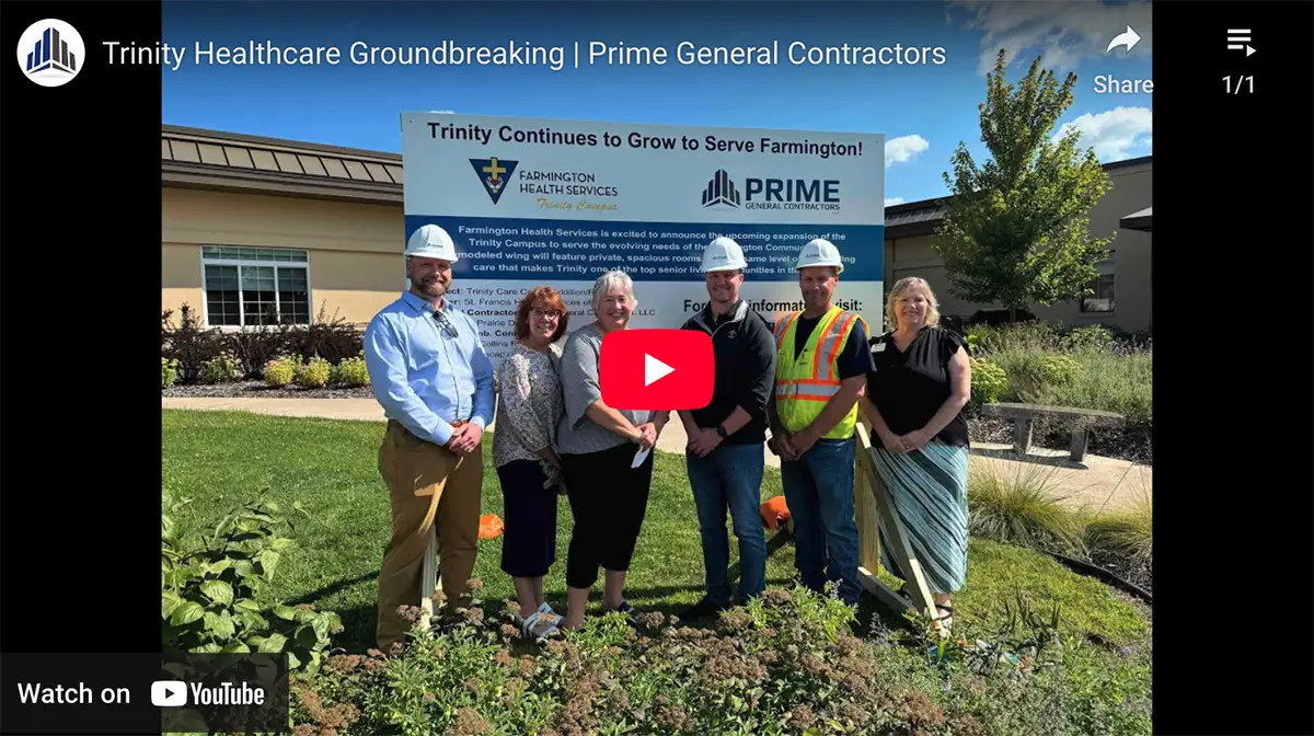 Six people stand in front of a building and a sign for Trinity Healthcare and Prime General Contractors at a Ground Breaking Ceremony.