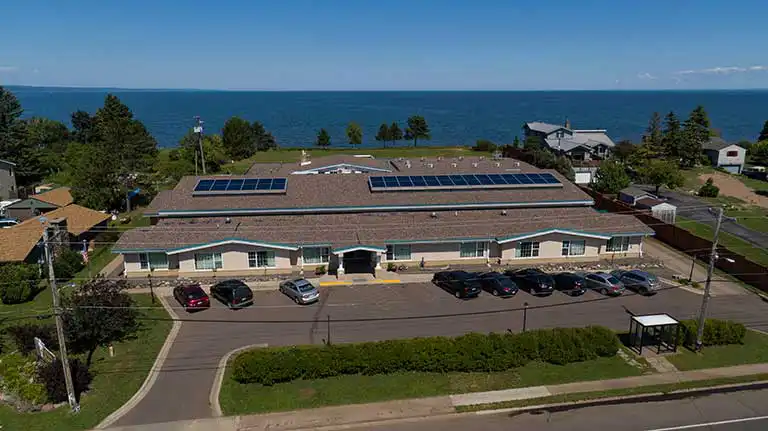 A single-story building with solar panels on the roof is surrounded by parked cars and greenery, with a large body of water visible in the background.