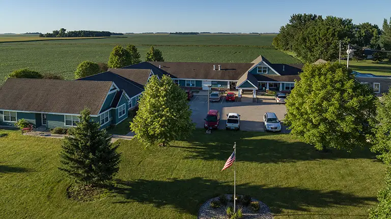Aerial view of a single-story residential building with multiple wings, a parking area with vehicles, trees, and an American flag on a grassy lawn, surrounded by farmland. Aerial view of a single-story residential building with multiple wings, a parking area with vehicles, trees, and an American flag on a grassy lawn, surrounded by farmland.