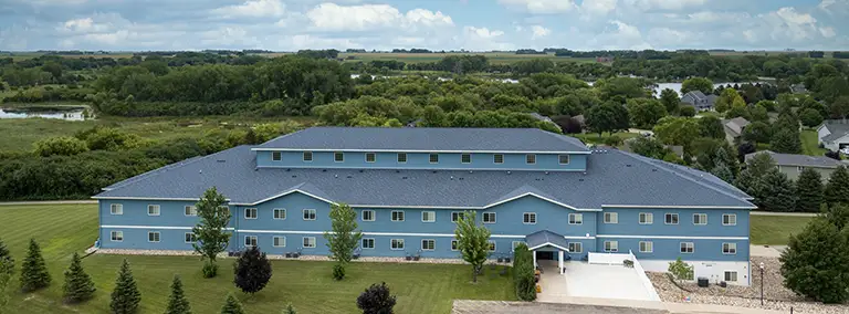 A large blue multi-story building with a dark roof is surrounded by lawns, trees, and distant countryside under a partly cloudy sky. A large blue multi-story building with a dark roof is surrounded by lawns, trees, and distant countryside under a partly cloudy sky.