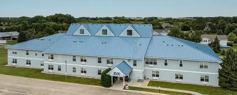 Two-story building with a blue metal roof, white exterior walls, and multiple dormer windows, surrounded by grass and residential neighborhood in the background.