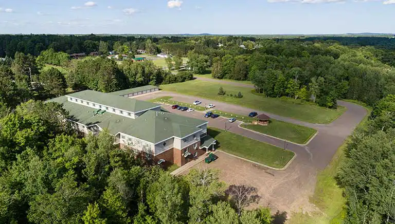 Aerial view of a large building surrounded by parking lots, trees, and greenery, with several parked cars and a forested landscape in the background.