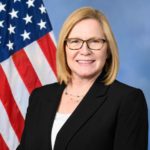 A woman in a black blazer and glasses stands in front of an American flag, smiling at the camera. A woman in a black blazer and glasses stands in front of an American flag, smiling at the camera.