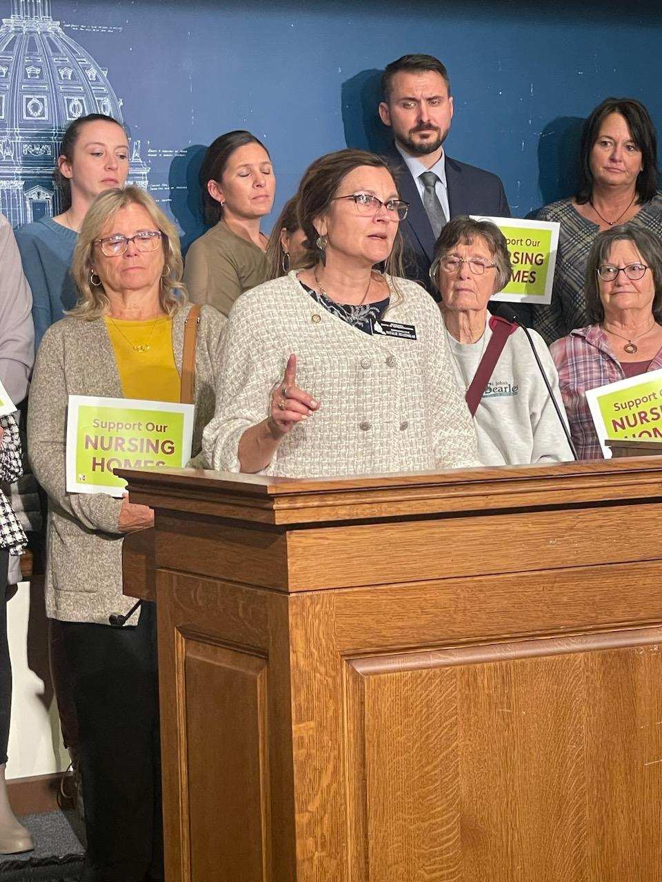 A woman speaks at a podium surrounded by people holding signs that read "Support Our Nursing Homes" and encourages everyone to Take Action Today.