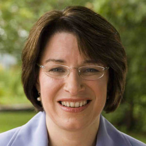 A woman with short brown hair and glasses is smiling outdoors, wearing a light-colored blazer. Trees and greenery are visible in the blurred background. A woman with short brown hair and glasses is smiling outdoors, wearing a light-colored blazer. Trees and greenery are visible in the blurred background.