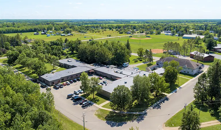 Aerial view of a building complex surrounded by trees, with a parking lot, nearby houses, open fields, and a baseball field in a rural area. Aerial view of a building complex surrounded by trees, with a parking lot, nearby houses, open fields, and a baseball field in a rural area.