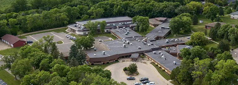 Aerial view of a large, single-story school complex with multiple wings, surrounded by dense trees and a parking area.