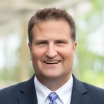 A man in a dark suit, light blue shirt, and patterned tie smiles at the camera with a blurred outdoor background, representing St. Francis Outpatient Therapy at St. Francis Health Services in Minnesota. A man in a dark suit, light blue shirt, and patterned tie smiles at the camera with a blurred outdoor background, representing St. Francis Outpatient Therapy at St. Francis Health Services in Minnesota.