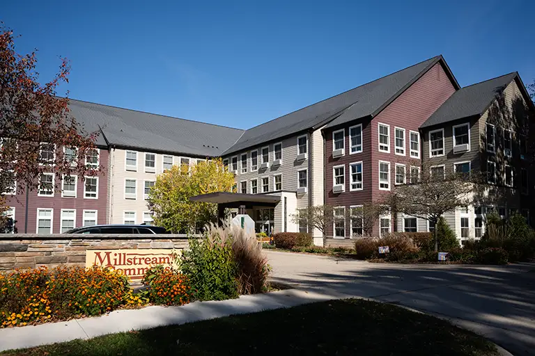 A three-story residential building with multiple windows and a peaked roof sits behind a sign reading "Millstream Commons" on a clear day.