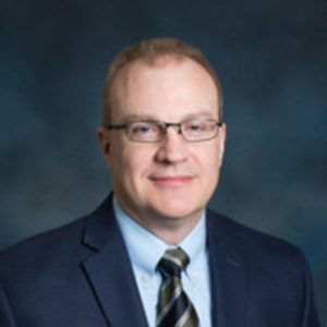 A man with short light brown hair and glasses, wearing a dark suit, light blue shirt, and striped tie, poses in front of a dark, blurred background representing St. Francis Outpatient Therapy.