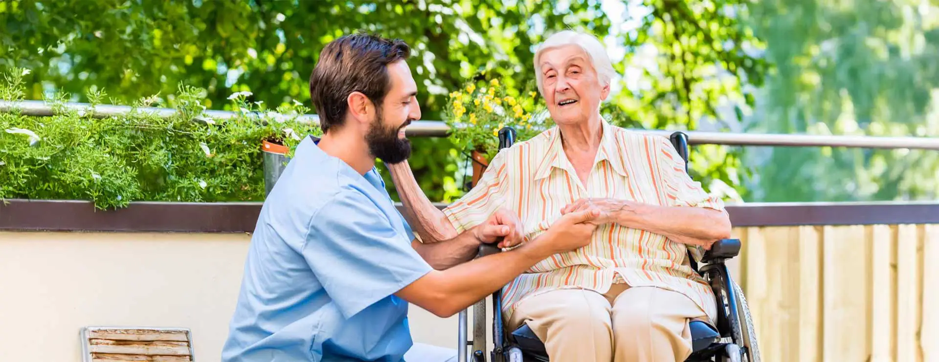 A caregiver kneels beside an elderly woman in a wheelchair, holding her hand and smiling, with greenery and sunlight in the background. A caregiver kneels beside an elderly woman in a wheelchair, holding her hand and smiling, with greenery and sunlight in the background.
