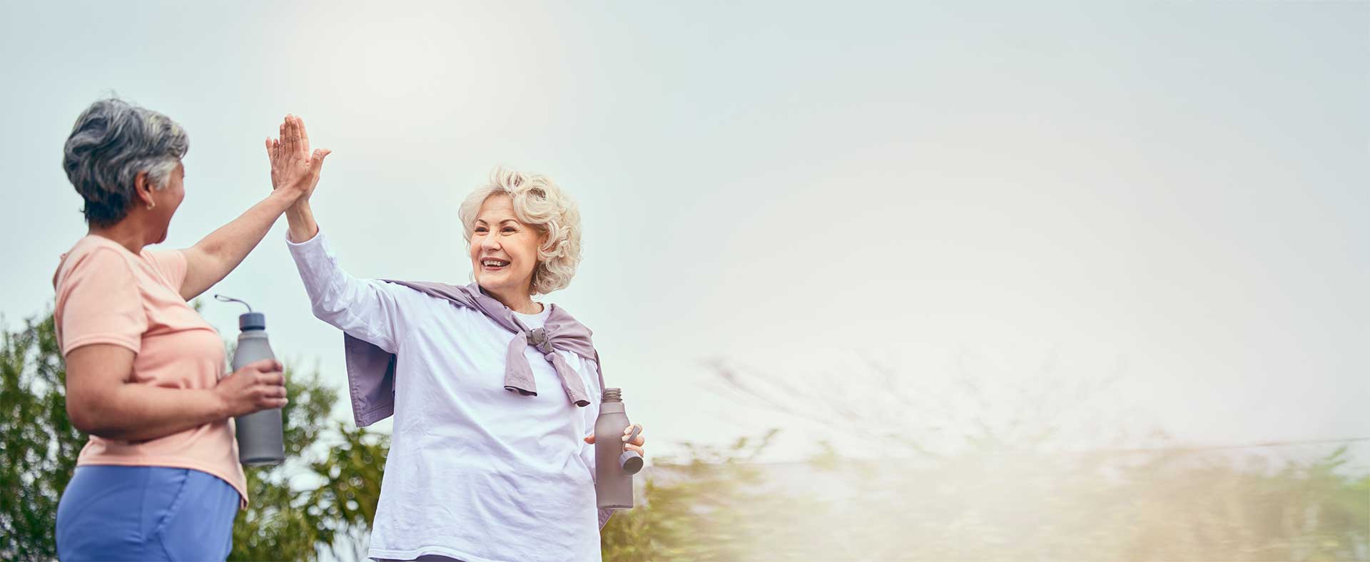 Two older women outdoors, smiling and giving each other a high-five while holding water bottles, with a bright sky in the background. Two older women outdoors, smiling and giving each other a high-five while holding water bottles, with a bright sky in the background.