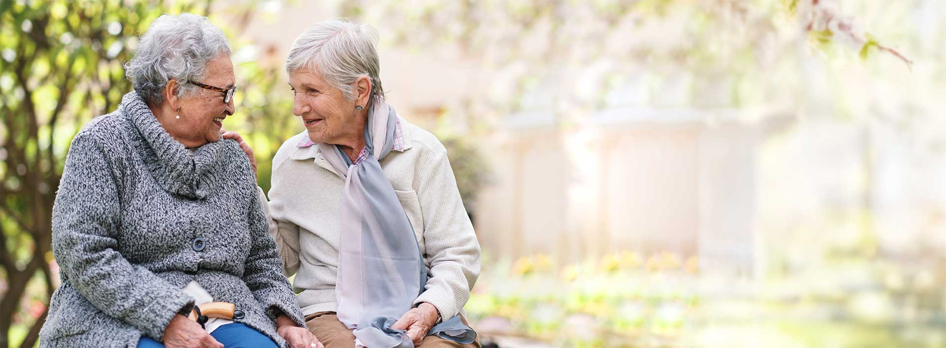 Two elderly women sit on a bench outdoors, smiling and talking to each other, with greenery and blurred background. Two elderly women sit on a bench outdoors, smiling and talking to each other, with greenery and blurred background.