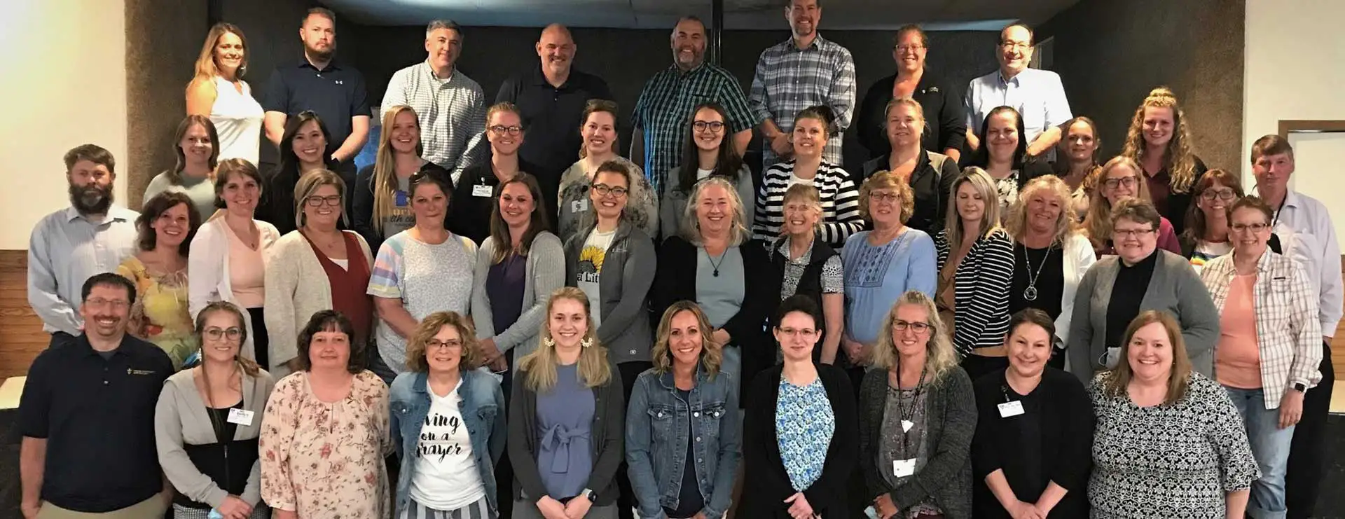 A large group of adults pose together indoors, standing and sitting in multiple rows, all facing the camera and smiling at St. Francis Outpatient Therapy.