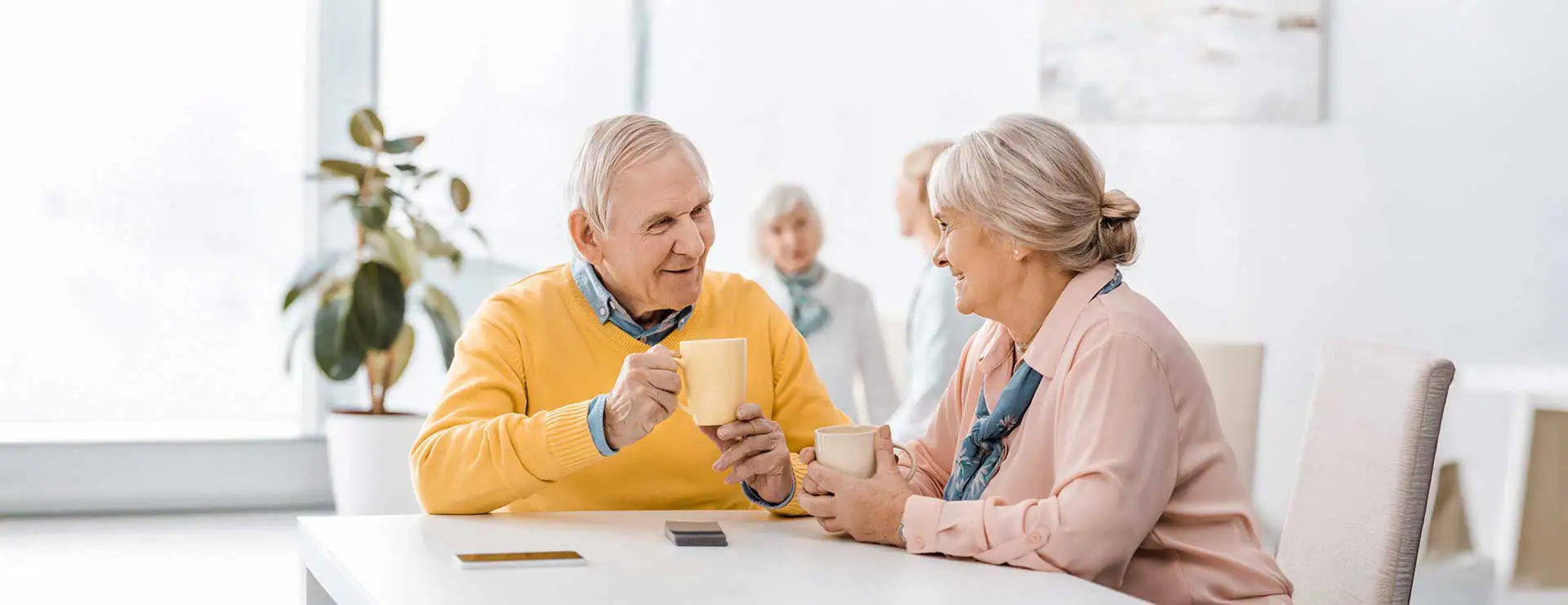 Two older adults sitting at a table, smiling and holding mugs, with two more people talking in the background in a bright room. Two older adults sitting at a table, smiling and holding mugs, with two more people talking in the background in a bright room.