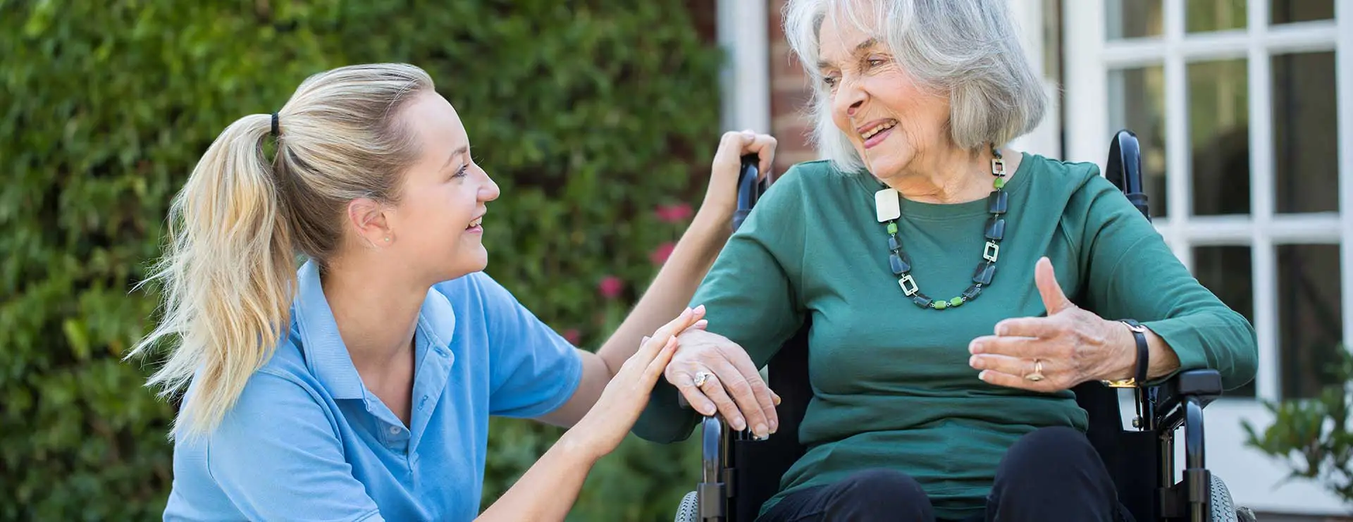 A caregiver in a blue shirt smiles and interacts with an elderly woman in a wheelchair outside near greenery. A caregiver in a blue shirt smiles and interacts with an elderly woman in a wheelchair outside near greenery.