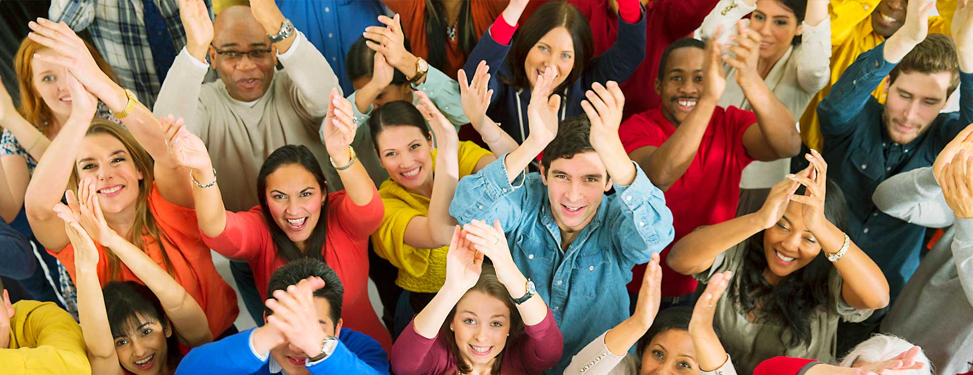 A diverse group of people stands close together, smiling and clapping while looking up toward the camera, celebrating the community spirit of St. Francis Outpatient Therapy.