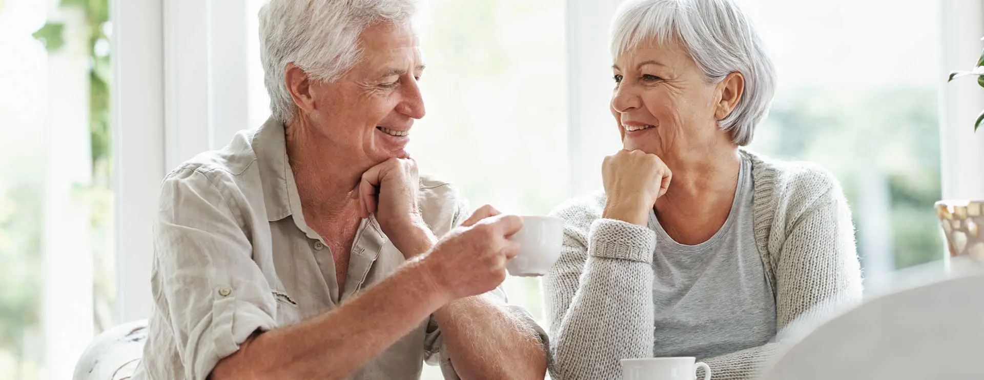 An older man and woman sit at a table indoors, smiling at each other while drinking from white mugs. An older man and woman sit at a table indoors, smiling at each other while drinking from white mugs.