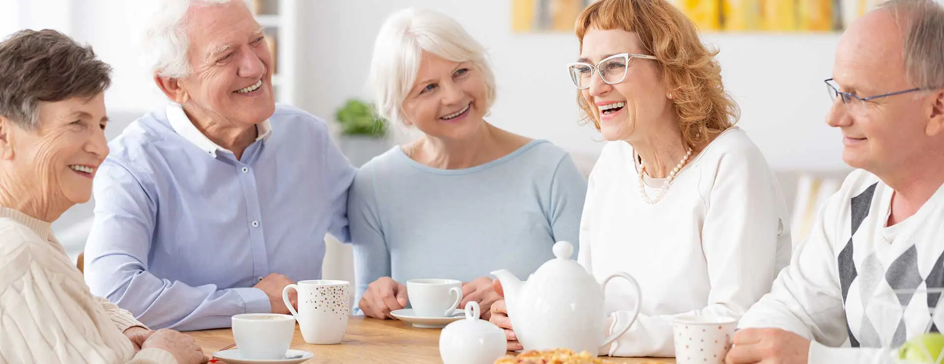 Five older adults sit at a table, drinking from mugs and smiling, with a teapot and pastries in front of them in a bright room.