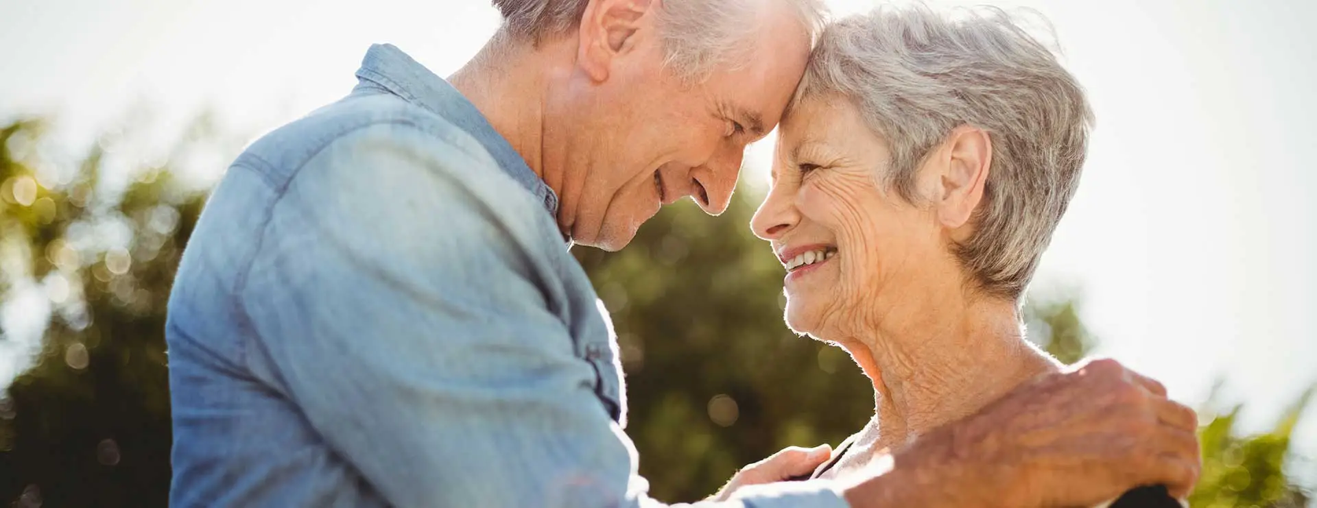 An older man and woman stand outdoors, touching foreheads and smiling at each other, with sunlight in the background. An older man and woman stand outdoors, touching foreheads and smiling at each other, with sunlight in the background.