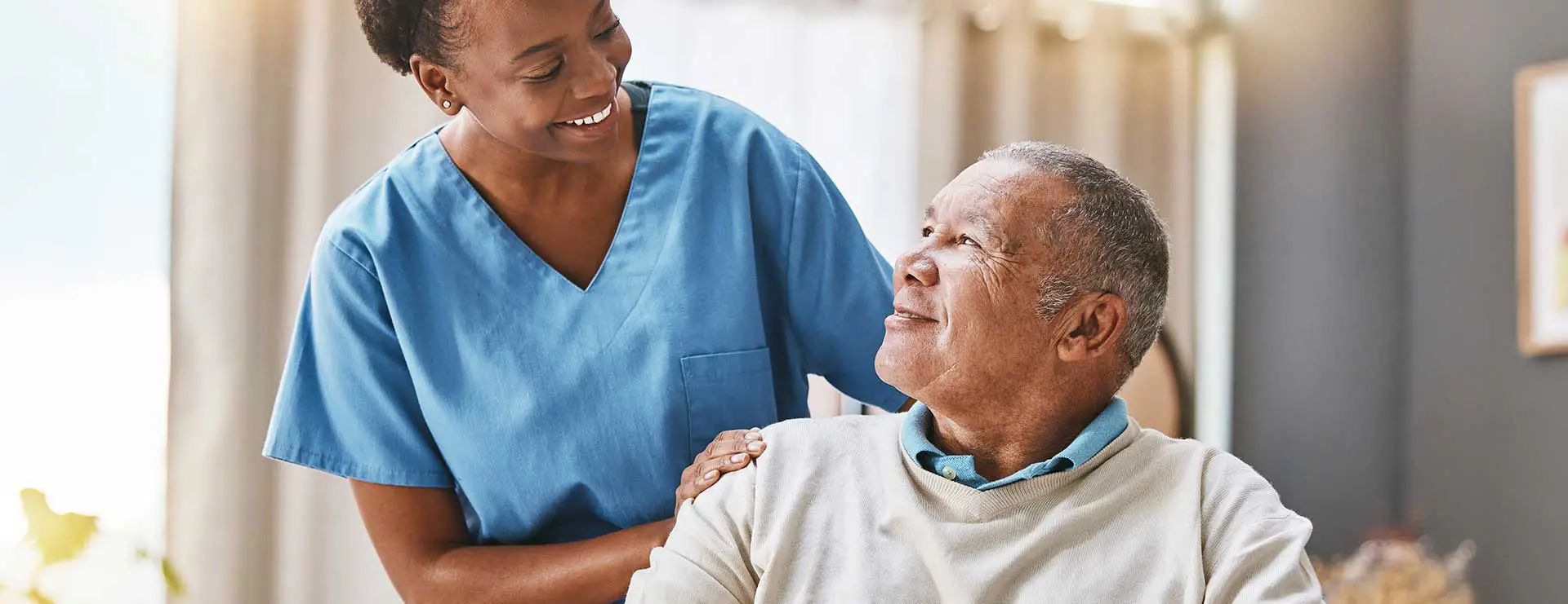 A nurse in blue scrubs smiles at an elderly man sitting and looking up at her in a well-lit room, suggesting a caring healthcare environment.