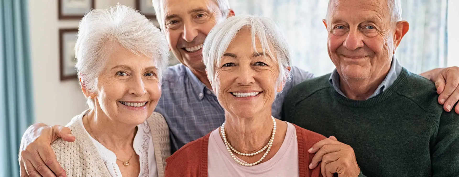 Four older adults stand closely together indoors, smiling at the camera. Two men and two women are visible, all with gray hair and wearing casual clothing. Four older adults stand closely together indoors, smiling at the camera. Two men and two women are visible, all with gray hair and wearing casual clothing.