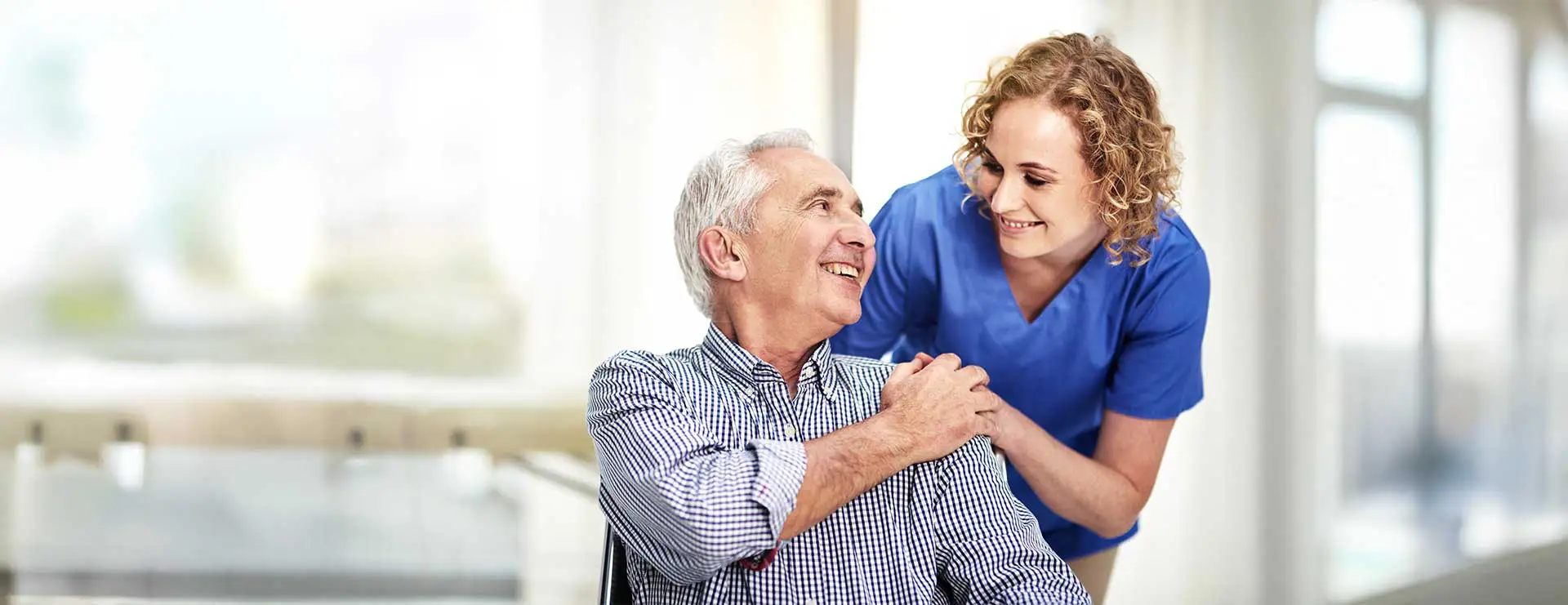A smiling elderly man in a wheelchair is being assisted by a female healthcare worker in blue scrubs, both looking at each other in a bright indoor setting. A smiling elderly man in a wheelchair is being assisted by a female healthcare worker in blue scrubs, both looking at each other in a bright indoor setting.