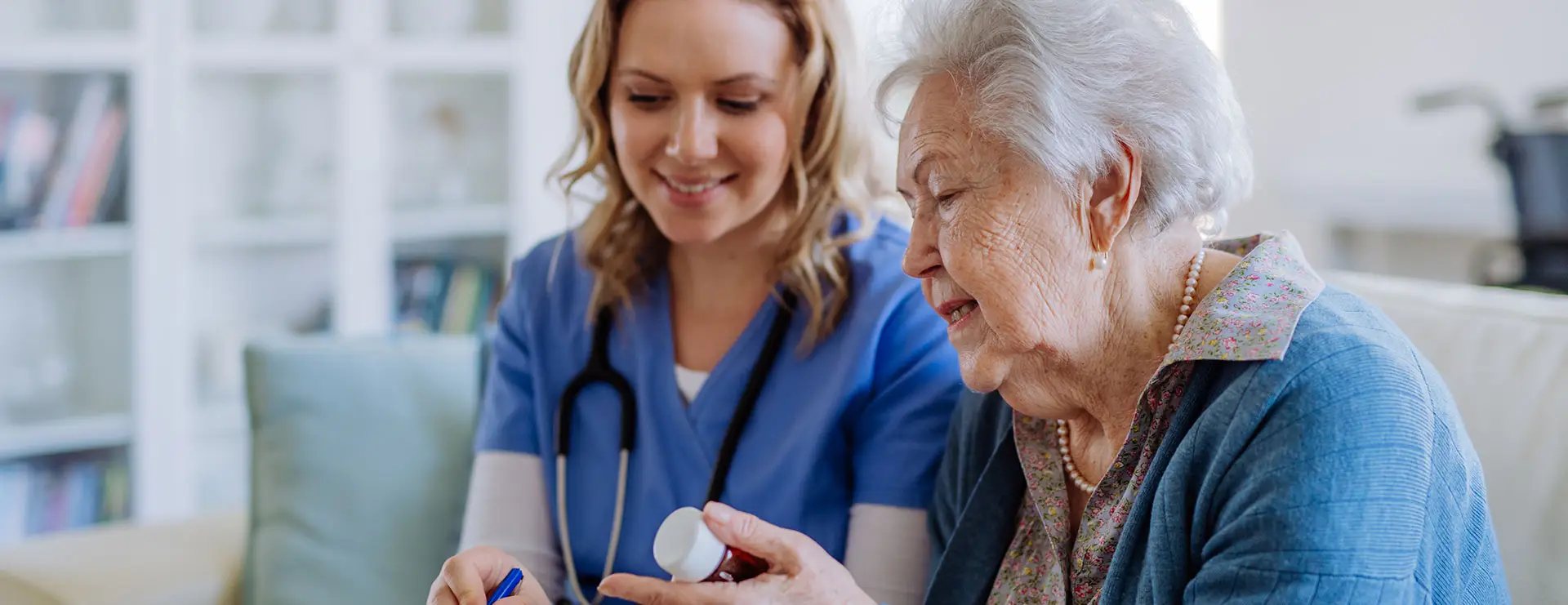 A nurse in blue scrubs sits with an elderly woman, discussing medication and writing notes, in a home setting. A nurse in blue scrubs sits with an elderly woman, discussing medication and writing notes, in a home setting.