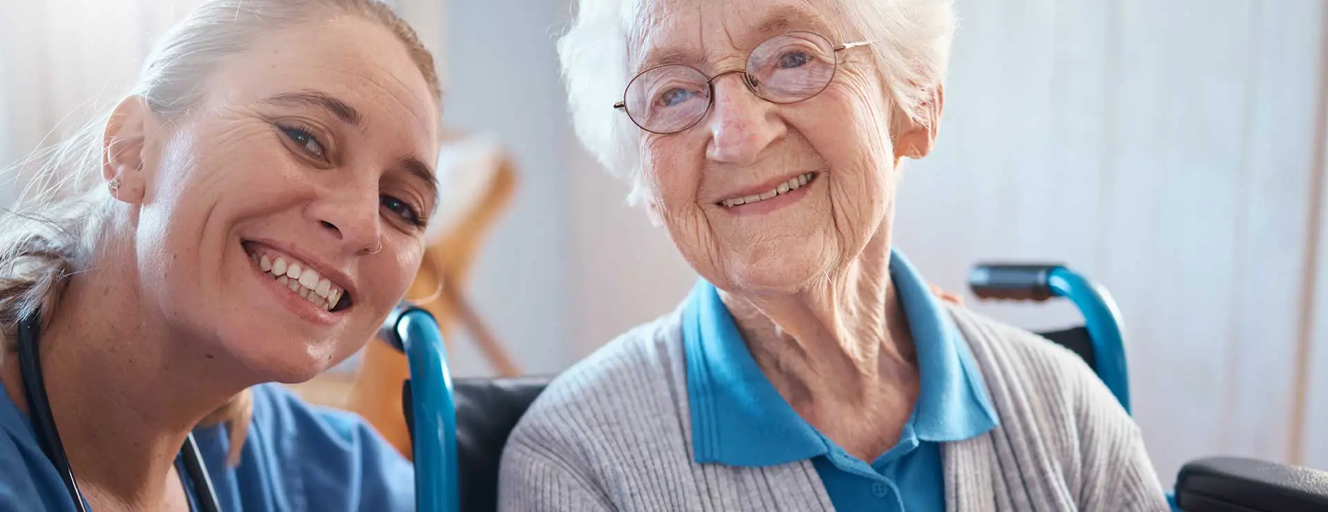 A younger woman and an older woman in a wheelchair smile at the camera in a brightly lit indoor setting. A younger woman and an older woman in a wheelchair smile at the camera in a brightly lit indoor setting.