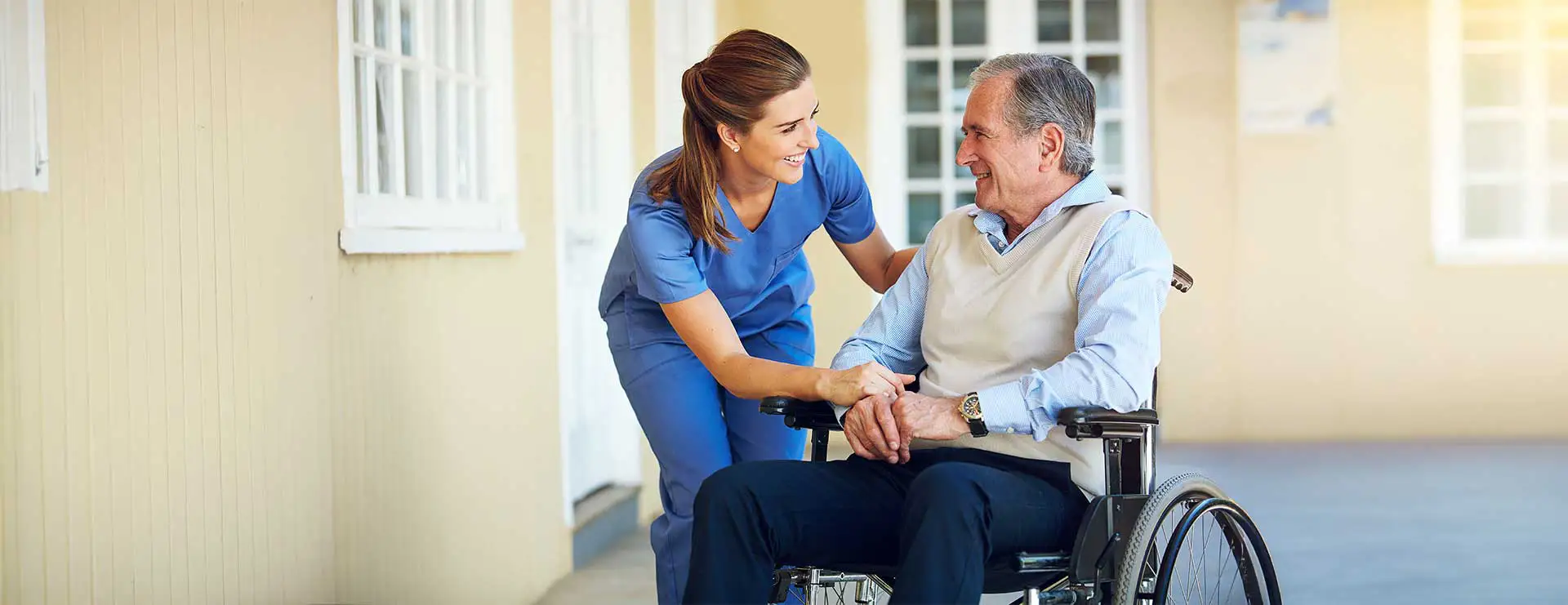 A nurse in blue scrubs smiles and talks with an older man sitting in a wheelchair outside a building, creating a friendly interaction. A nurse in blue scrubs smiles and talks with an older man sitting in a wheelchair outside a building, creating a friendly interaction.