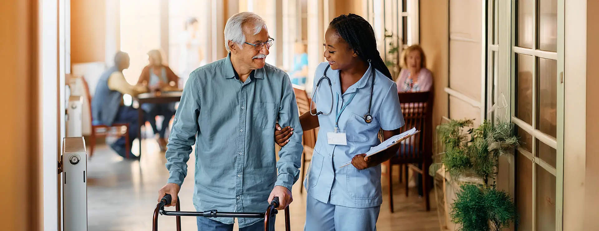 A nurse from St. Francis Health Services assists an elderly man using a walker as they walk down a hallway in a care facility. A nurse from St. Francis Health Services assists an elderly man using a walker as they walk down a hallway in a care facility.