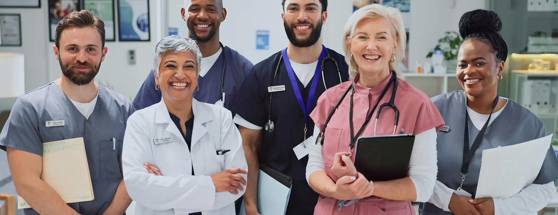 Six healthcare professionals stand together in a clinical setting, smiling at the camera; some hold clipboards, wearing scrubs and lab coats.