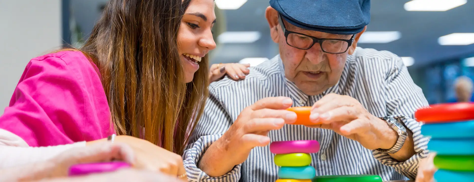 A young woman and an elderly man stack colorful plastic rings together at a table in a bright indoor setting at St. Francis Outpatient Therapy in Minnesota. A young woman and an elderly man stack colorful plastic rings together at a table in a bright indoor setting at St. Francis Outpatient Therapy in Minnesota.