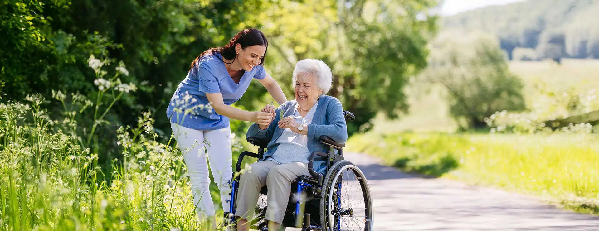 A caregiver assists an elderly woman in a wheelchair while they are outside on a sunny path surrounded by greenery. A caregiver assists an elderly woman in a wheelchair while they are outside on a sunny path surrounded by greenery.