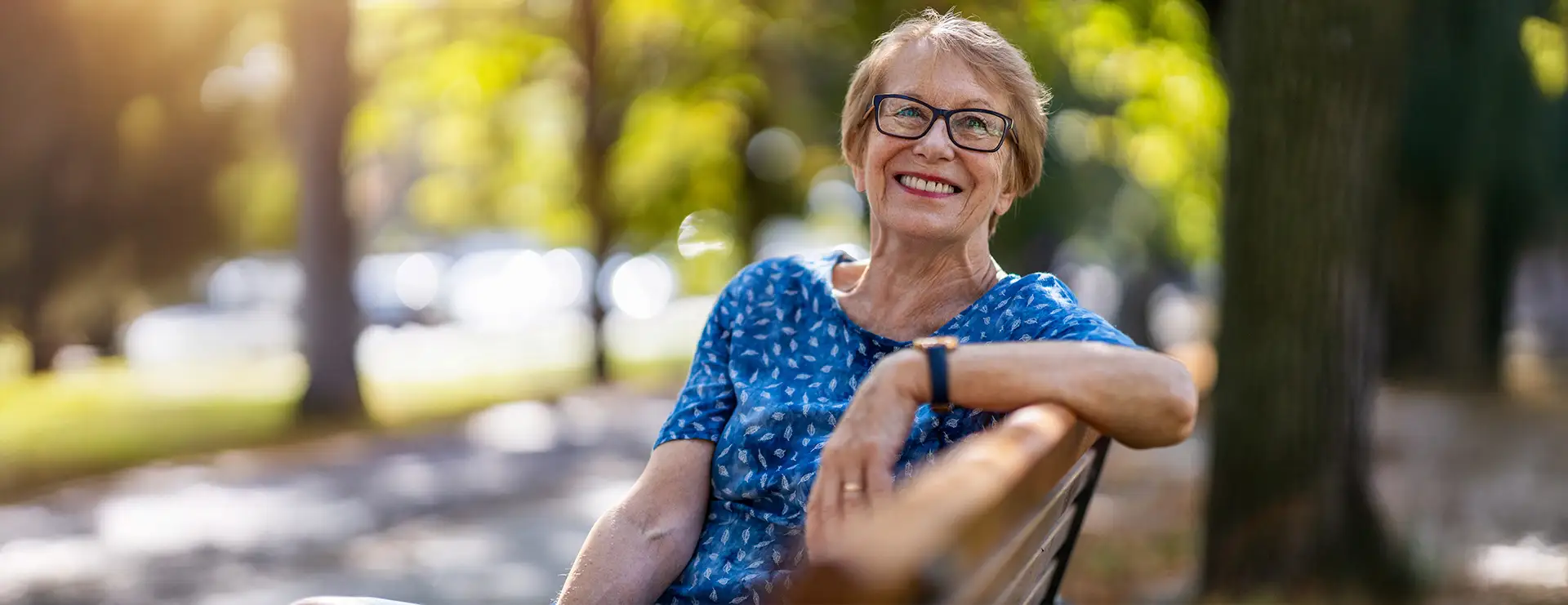 An older woman wearing glasses and a blue patterned shirt sits on a bench outdoors in Farmington, smiling, with sunlight and trees in the background—reflecting the warmth of quality health services in her community.