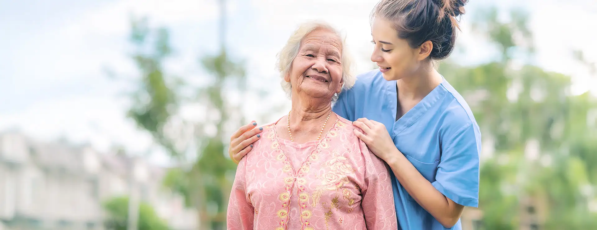 A young caregiver in blue scrubs stands outdoors with her arms around an elderly woman in a pink dress, both smiling against a blurred background of trees and buildings at Trinity Care Center. A young caregiver in blue scrubs stands outdoors with her arms around an elderly woman in a pink dress, both smiling against a blurred background of trees and buildings at Trinity Care Center.