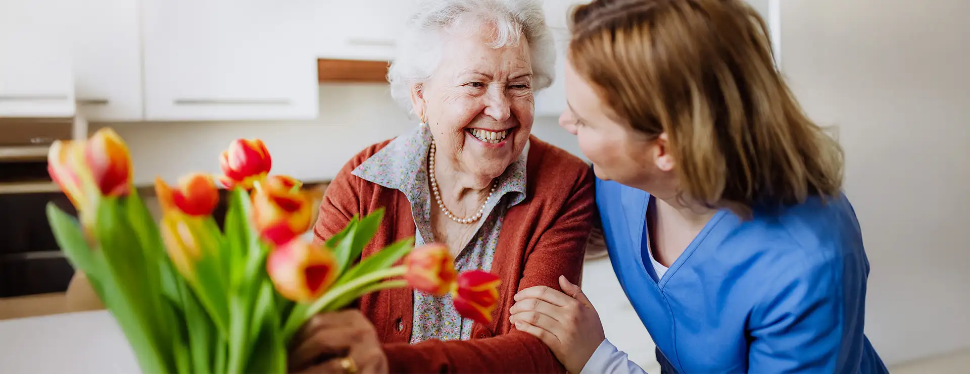 An elderly woman smiles while sitting at a table with a bouquet of tulips, as a Trinity Terrace caregiver in blue scrubs holds her arm and smiles back. An elderly woman smiles while sitting at a table with a bouquet of tulips, as a Trinity Terrace caregiver in blue scrubs holds her arm and smiles back.