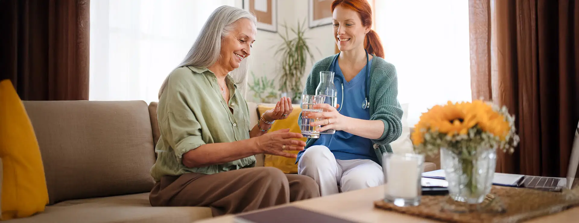 A Trinity Suites caregiver pours a glass of water for an older woman sitting on a couch in a well-lit living room with yellow accents and sunflowers, ensuring compassionate Memory Care. A Trinity Suites caregiver pours a glass of water for an older woman sitting on a couch in a well-lit living room with yellow accents and sunflowers, ensuring compassionate Memory Care.