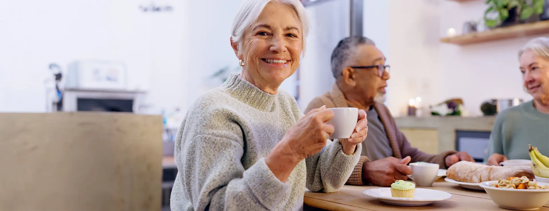 Older adults sit at a kitchen table enjoying hot drinks and pastries, with one woman smiling at the camera while holding a cup, sharing a warm moment supported by Koochiching Health Services. Older adults sit at a kitchen table enjoying hot drinks and pastries, with one woman smiling at the camera while holding a cup, sharing a warm moment supported by Koochiching Health Services.