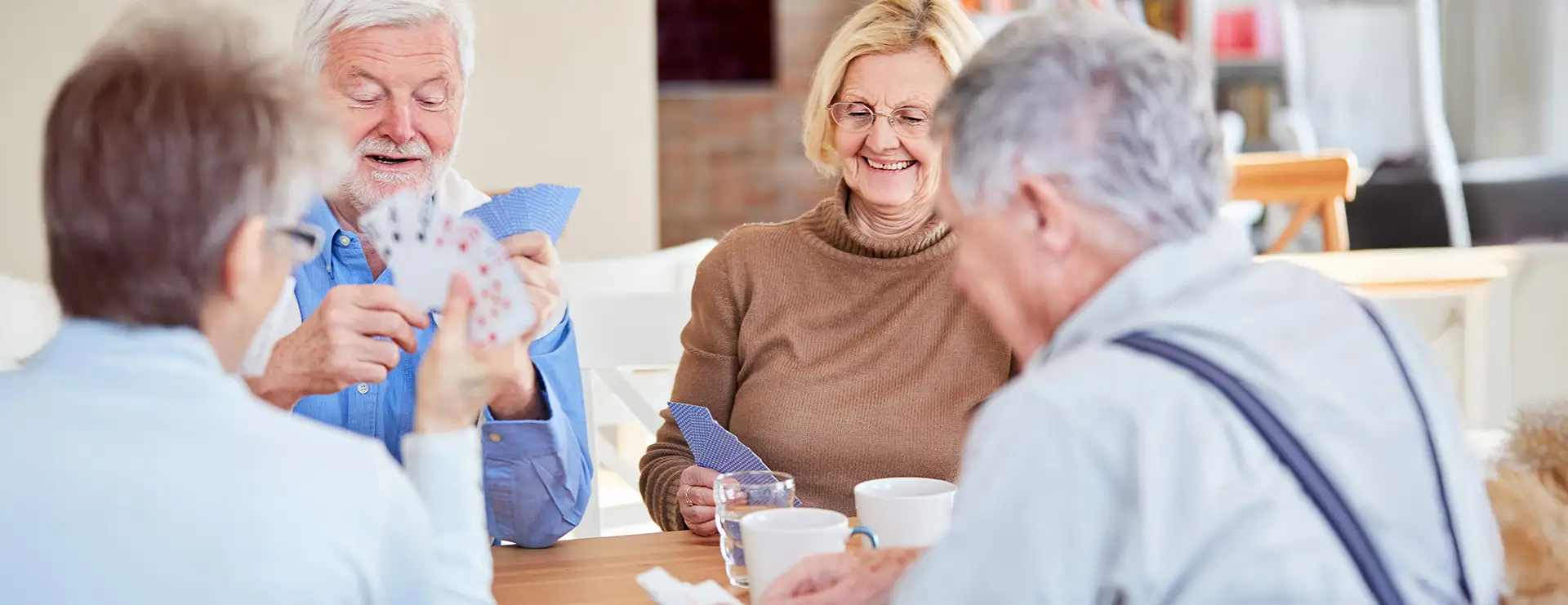Four older adults sit around a table at Skyview Court Apartments, playing cards and drinking from mugs. Two people are smiling while one person holds a spread of cards, capturing a moment of camaraderie and fun.