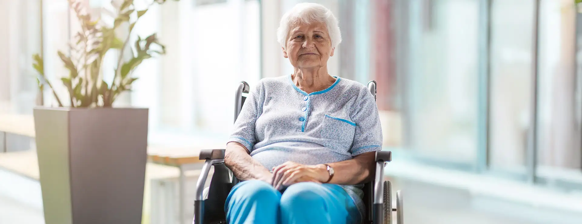 An elderly woman with short gray hair sits in a wheelchair indoors, wearing a light blue shirt and blue pants, with a potted plant visible nearby. An elderly woman with short gray hair sits in a wheelchair indoors, wearing a light blue shirt and blue pants, with a potted plant visible nearby.