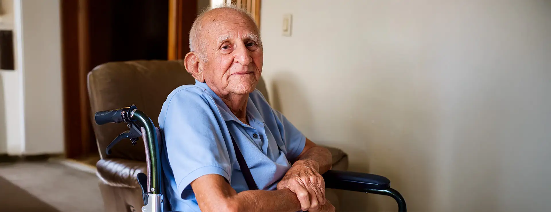 An elderly man wearing a light blue shirt sits in a wheelchair indoors at Little Falls Care Center, looking at the camera with a neutral expression. An elderly man wearing a light blue shirt sits in a wheelchair indoors at Little Falls Care Center, looking at the camera with a neutral expression.