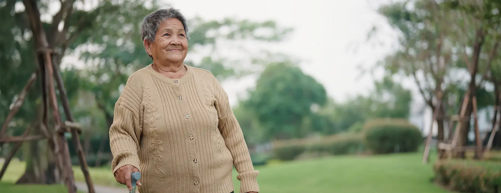 An older woman wearing a beige sweater stands outdoors in a park, holding a cane and smiling, with trees and greenery in the background—showcasing the active lifestyle supported by Morris Health Services. An older woman wearing a beige sweater stands outdoors in a park, holding a cane and smiling, with trees and greenery in the background—showcasing the active lifestyle supported by Morris Health Services.